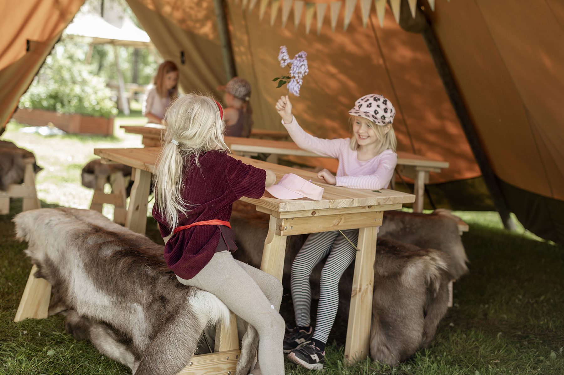 Børn sidder om et træbord under en tipi og leger med blomster