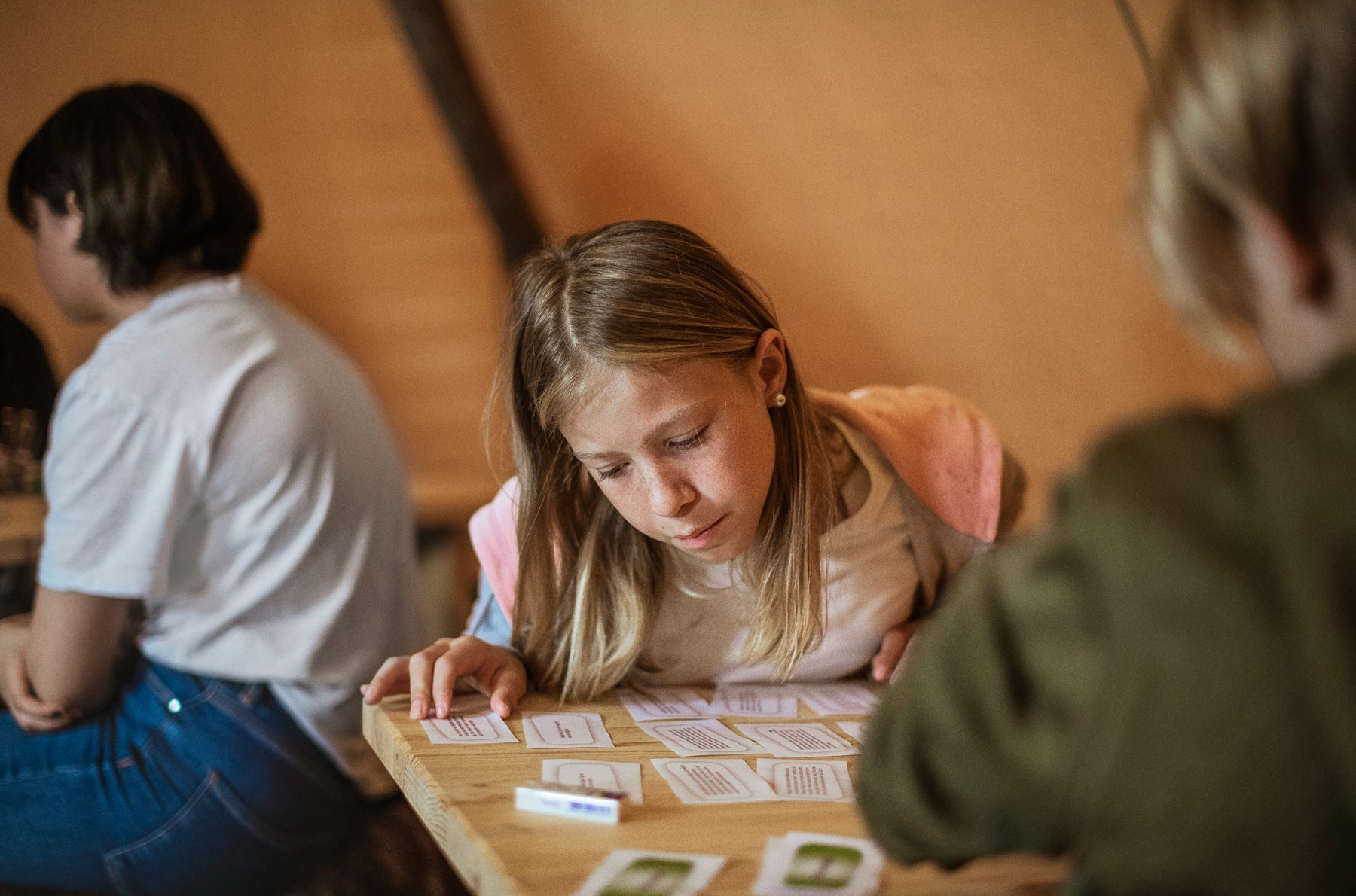 Barn koncentrerer sig om et brætspil ved et træbord under teltdugen
